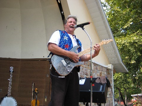 A photo of Dave Stamm from the band Lucky Afternoon plays guitar and sings into the microphone he has setup on the Knoebels Hawaiian Bandshell.