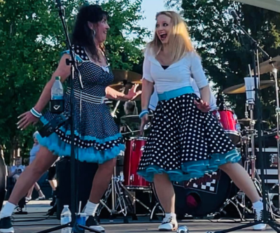 Two lead female singers of the Main Street Cruisers pose looking at each other mid-song. They're wearing nostalgic black with white spot mid-century dresses with blue accents. 